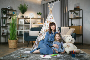 Pleasant caucasian mother gently embracing her daughter while sitting together on floor near toy wigwam and smiling on camera. Little kid studying to draw in playful manner. © sofiko14