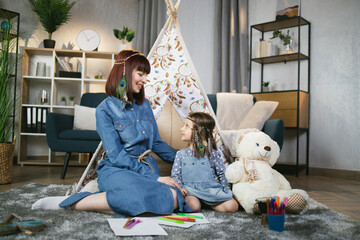 Charming caucasian woman and cute little child sitting on floor near teepee tent and smiling to...