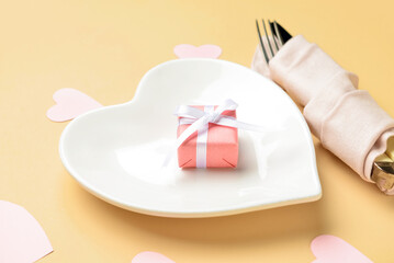 Beautiful heart-shaped plate with gift and cutlery on beige background, closeup