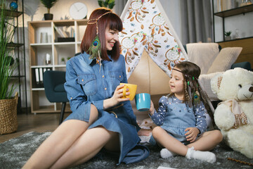 Smiling woman drinking tea with her cute daughter while playing near toy wigwam at home. Creative time spending of caucasian family. © sofiko14