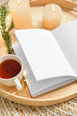 Tray with cup of tea, opened book and burning candles on wicker surface, closeup