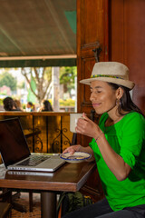 Latin woman wearing a hat, sitting in a old coffee shop working on her laptop holding a cup of coffee