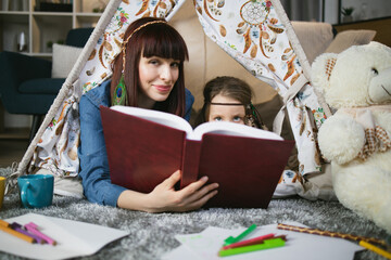Attractive caucasian woman lying with cute female child inside colorful toy wigwam and reading book together. Mother and daughter studying in playful manner at home.