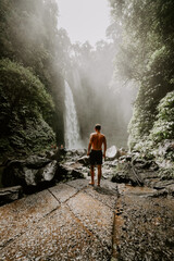 Obraz premium A young male traveller walking amongst green lush tropical forest towards a high waterfall in Bali jungle 