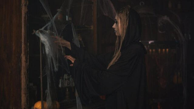 Halloween Witch take potion from shelf with spider web, candle on background, magic everywhere. Dark witch dressed black cloak selects ingredients for making love potion or poison Handheld 