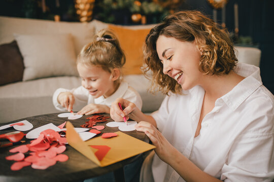 Mother and daughter making Valentine's day cards using color paper, scissors and pencil, sitting by the table in cozy room