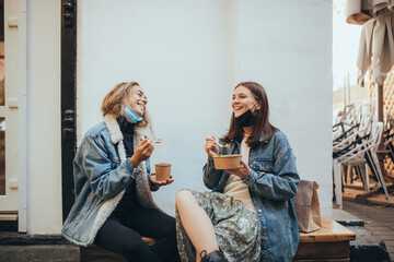 Two young women female friends wearing protective face mask sitting outdoors eating takeaway food,...