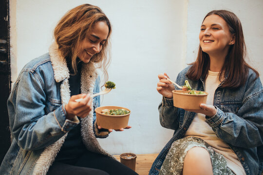 Two young women female friends sitting outdoors eating takeaway food, laughing and having fun. Food delivery and takeout.