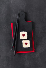 Two mousse creamy white chocolate cakes with strawberry jelly hearts, in the form of square sweet sushi, sprinkled with coconut chips, on a serving board on dark background. Top view.  Valentine's Day