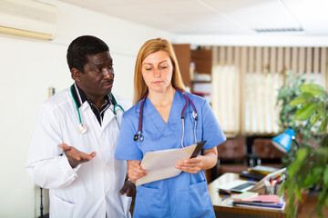 Two multinational doctors working at the clinic discuss work issues while standing in a well-lit...