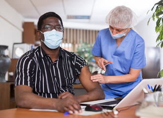 Professional male nurse injecting vaccine in shoulder of male patient during covid vaccination in office