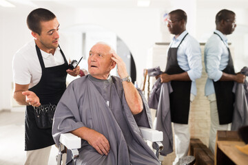 Obraz premium Portrait of shocked elderly man sitting in barber chair with confused young hairdresser behind him