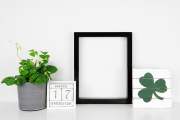 Mock up black frame with St Patricks Day decor on a white shelf. Shamrock plant, shabby chic wood calendar and sign. Portrait frame against a white wall. Copy space.