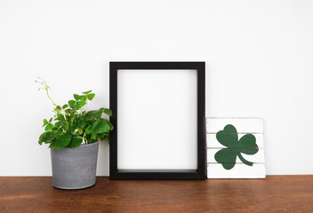 Mock up black frame with St Patricks Day decor on a wood shelf. Shamrock plant and shabby chic wood sign. Portrait frame against a white wall. Copy space.