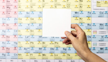 Woman hand holding blank white paper cardboard sheet against the blurred periodic chart of the...
