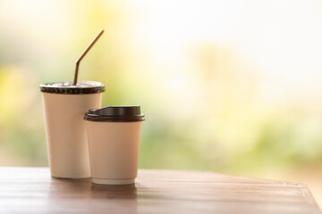 Closeup of disposable take away paper cup of hot and iced coffee on wooden table with green nature background.