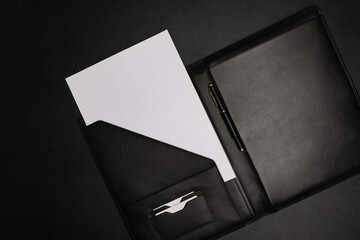 Black leather file folder agenda organizer opened with white blank paper sheet and pen on black background with copy space