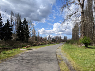 View of a paved biking and walking trail along a river on a bright, sunny day with thick white clouds in the sky