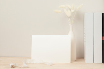 Blank paper card, greeting card mock up. decoration with dried Bunny Tail, Front view, Beautiful Bunny Tail grass in vase and white book on wood table and beige cement wall background