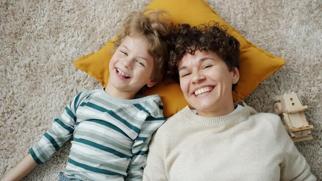 Top view slow motion of happy young mother and son laughing looking at camera lying on floor at home. Positive emotion and family concept.