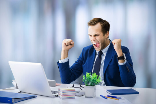 Very happy business man in blue suit, working with laptop computer at modern office. Excited business man rising hands up with clenching fists - success in job concept image.