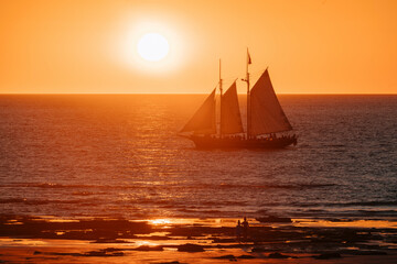 The Willie Pearl Lugger cruises along the calm waters of Cable Beach, Broome, in the north of Western Australia during a vibrant glowing sunset while two young children watch from the shore.