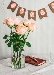 Bouquet of delicate roses in a glass vase, a leather notebook and easter paper handmade garland on a table in a bright room