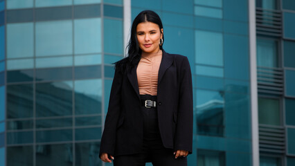 Young hispanic businesswoman employee company standing confidently outdoors on background office building looking at camera posing in successful pose feels satisfied with career growth promotion