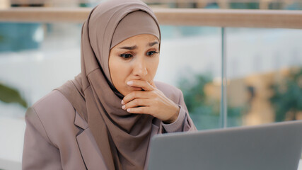 Sad muslim girl student in hijab looking at laptop screen reading email shocked by bad news...