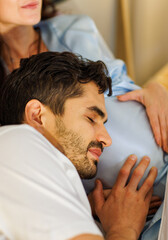 Delighted father tenderly lie down on belly of beloved pregnant wife on bed to feel and hear unborn baby kicking in stomach