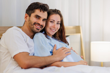 Fototapeta premium Cheerful husband enjoy embracing pregnant wife and happy to tenderly fondle beloved belly of unborn baby and expecting warm family during prenatal care of maternity at romantic bedroom