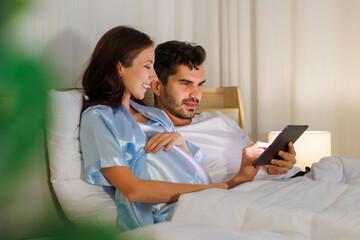 Cheerful pregnant mother enjoy kidding play with unborn baby via online video on tablet computer while father smiling in a bed room