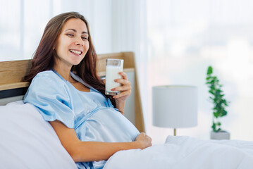 Cheeful pregnant woman smile as enjoy drinking glass of milk beverage in morning while resting on bed with lovely bear doll