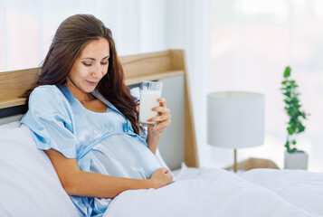 Cheeful pregnant woman smile as enjoy drinking glass of milk beverage in morning while resting on bed with lovely bear doll