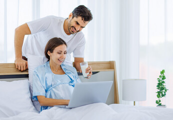 Lovely pregnant wife tenderly touch beloved belly of unborn baby while lounging on bed with smile and happy as enjoy watching entertainment media on computer