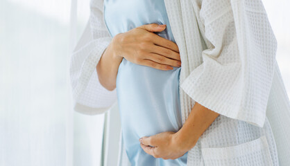 Cheerful pregnant woman on night gown laugh with happy and fun to hold belly of unborn baby while standing near window curtains of living room for refreshing
