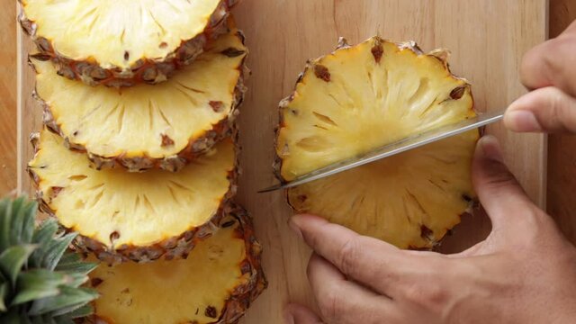 CU Close-up of Man's hand slicing pineapple with a knife on a wooden cutting board. 
