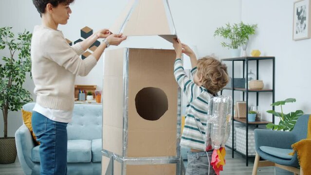 Slow motion of young woman and kid wearing astronaut costume making cardboard spaceship at home. Childhood and parenthood concept.