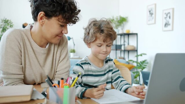 Mother teaching little boy talking looking at laptop screen writing in notebook at home. Family education and homeschooling concept.