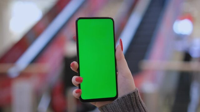 Hands of a young woman holding a smartphone in a shopping mall. A young woman is holding a phone in a shopping mall in front of an escalator with people. Close up