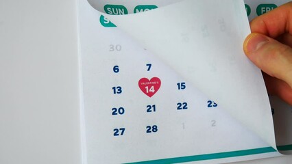 Close-up of a male hand flipping through a February page of the wall calendar followed by the next one with the date of Valentine's Day marked with a pink heart shaped symbol - Powered by Adobe