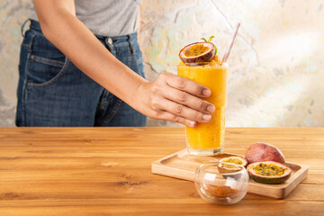 Asian woman taking a glass of passion fruit smoothie on a wooden tray with passion fruit on the table over abstract colorful wall background backdrop. Eatable recipe healthy vegan fruit shake concept