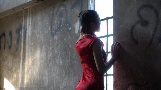 Chinese girl in red chongseum chinese traditional clothing in a rustic building standing to windows light.