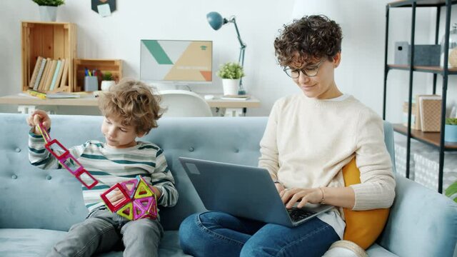 Slow motion of young woman working with laptop typing and playing with child multitasking at home. Family life and motherhood concept.