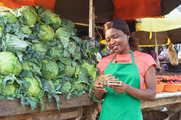 Happy African business woman or female trader wearing a green apron and counting Naira notes while...