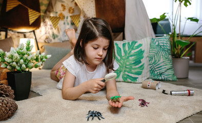 Girl playing observing toy bugs with a magnifying glass lying on the carpet