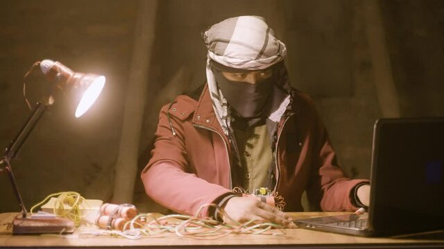 Gangster with face cover secretly making Bomb by seeing online videos on laptop at abandoned place - concept of planning for crime and technology used for illegal or unlawful activities.