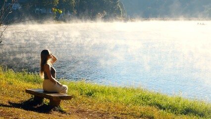 Fototapeta premium Travel woman relax on nature near misty lake. Female traveler sitting on bench, enjoy fog morning on river. Calmness, relaxation, meditation, freedom, beauty of nature concept