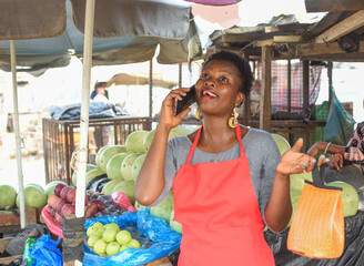 African woman or female trader making calling with a smart phone as she stands at her stall in a...