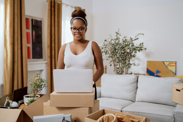 Smiling woman in glasses is standing in middle of an apartment during move, around her cardboard...